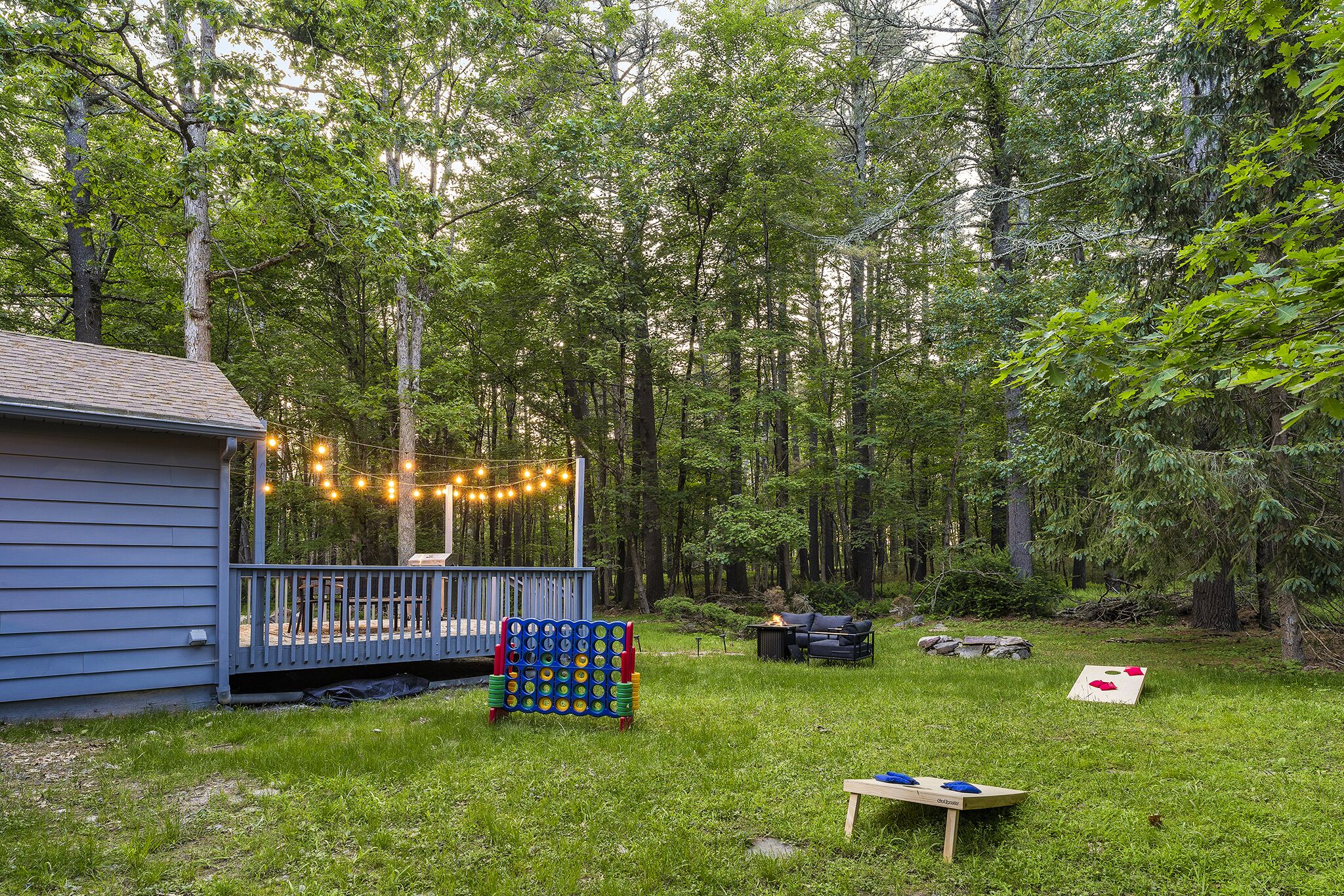 A backyard with a wooden deck, string lights, a blue house, and a grassy area with a table and chairs.