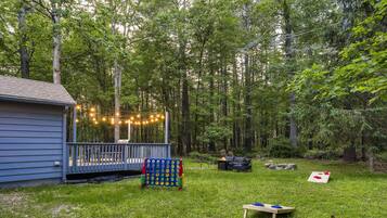 A backyard with a wooden deck, string lights, a blue house, and a grassy area with a table and chairs.