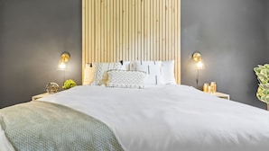 A modern bedroom with a wooden headboard, a large bed with patterned bedding, two wall-mounted lights, and a bedside table with a plant and candles. - Retro Countryside Cabin | Cozy Upstate NY Private Retreat! (Eldred)