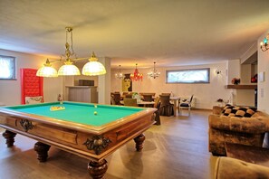 A spacious living room with a pool table, a red cushioned chair, and a sofa. There are multiple dining tables and chairs, a chandelier, and a fireplace.