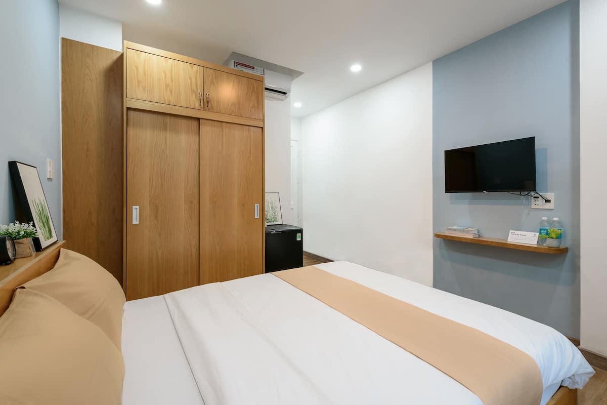 A bedroom with a bed, wooden wardrobe, a small black appliance, a mounted TV, and a shelf with items.