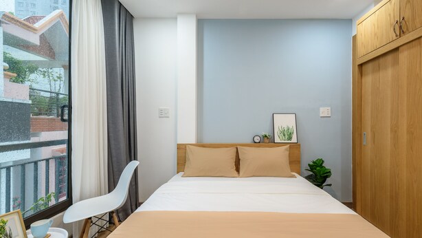 A modern bedroom with a large bed, a bedside table with a cup and a picture frame, a chair, a wardrobe, and a window with curtains.