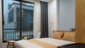 A hotel room with a bed, a chair, a small table, a window with curtains, and a view of buildings outside.