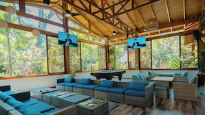 A spacious indoor area with wicker furniture, a pool table, and large windows offering a view of greenery.