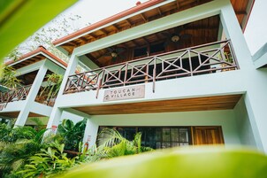 A multi-story building with balconies, wooden beams, and a sign reading "TOUCAN VILLAGE".
