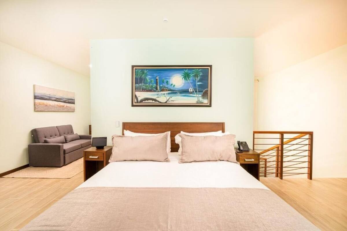 A hotel room with a large bed, a nightstand, a desk, a chair, a sofa, a painting, and a staircase.