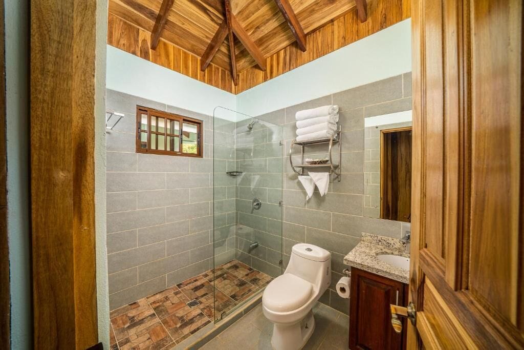 A bathroom with a shower, toilet, and sink, featuring wooden ceiling beams and tiled walls.