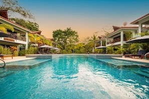 Piscina con sillones de descanso y sombrillas, rodeada de vegetación y edificios.