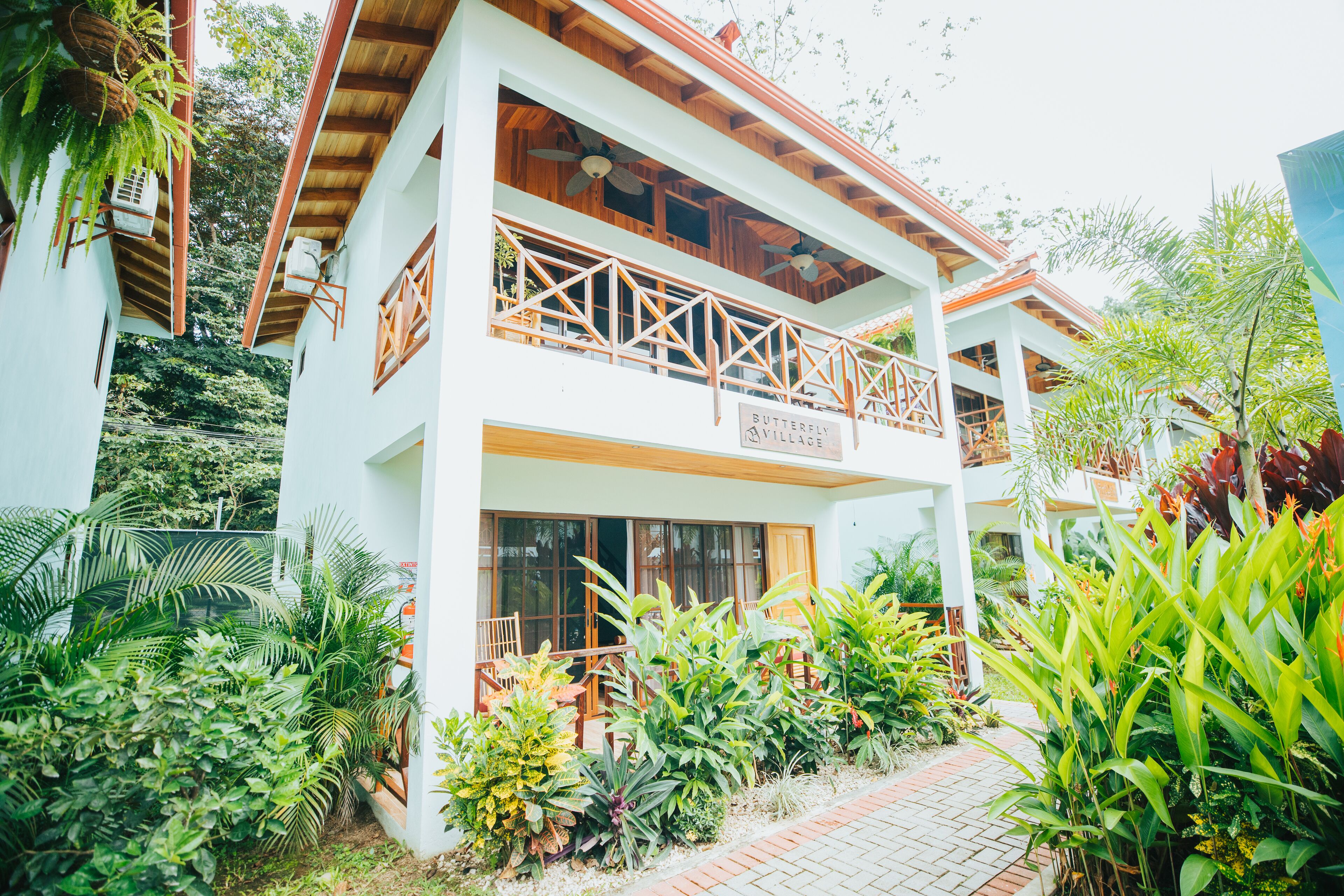 Um edifício de dois andares com uma varanda, rodeado por vegetação exuberante e uma placa com a inscrição 'Butterfly Village'.