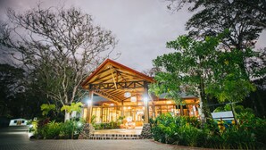 A wooden lodge with a covered entrance, surrounded by lush greenery and a paved driveway.