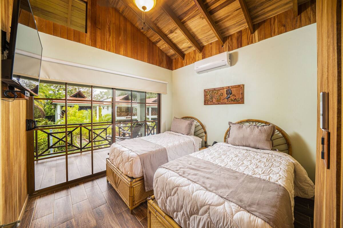 A bedroom with two beds, wooden flooring, a large window, and an air conditioning unit.