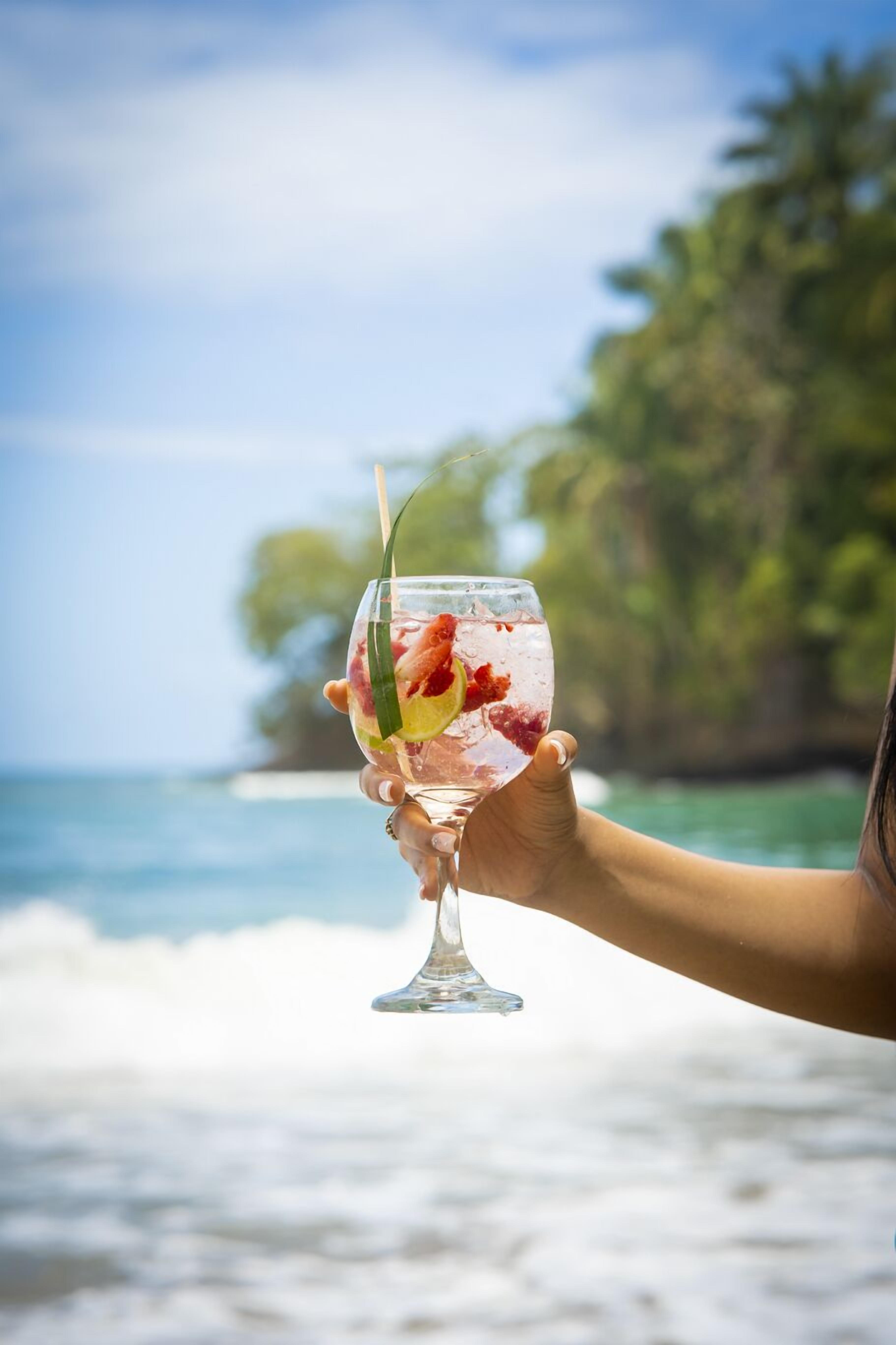 A hand holding a cocktail glass with fruit and a straw, against a beach backdrop with waves and greenery.