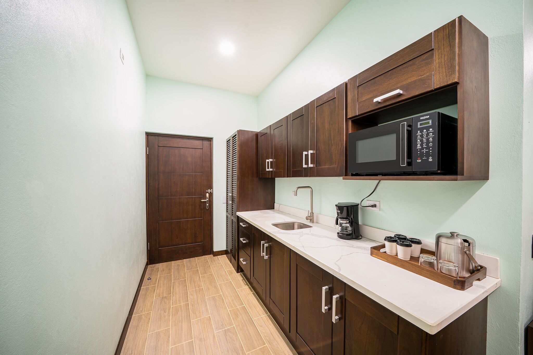 A compact kitchen with a microwave, coffee maker, and a tray with cups and a thermos.