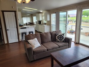 A living room with a brown sofa, a coffee table, and a view of a kitchen and outdoor patio. - 6th Hole Loop in Desert Aire (Mattawa)