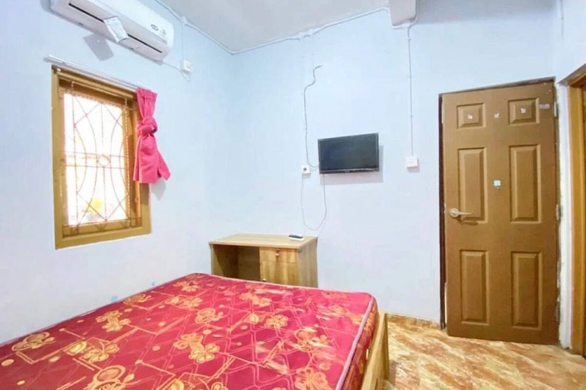 Une chambre à coucher comprenant un lit, un téléviseur, un climatiseur, une fenêtre avec un rideau et une porte.