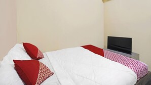 Een enkel bed met wit en rood beddengoed, een flatscreen televisie op een nachtkastje en een wandairconditioner.