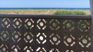 A view through an arched window with a patterned metal grille, overlooking a beach with a clear sky.