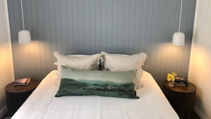 A neatly made bed with white linens and two pillows, one with a landscape design. There are two bedside tables, each with a lamp and a book. The room has a simple, clean design with a grey wall and a hanging light fixture. - Centrally located "Bargara Beach Cottage" (Bargara)