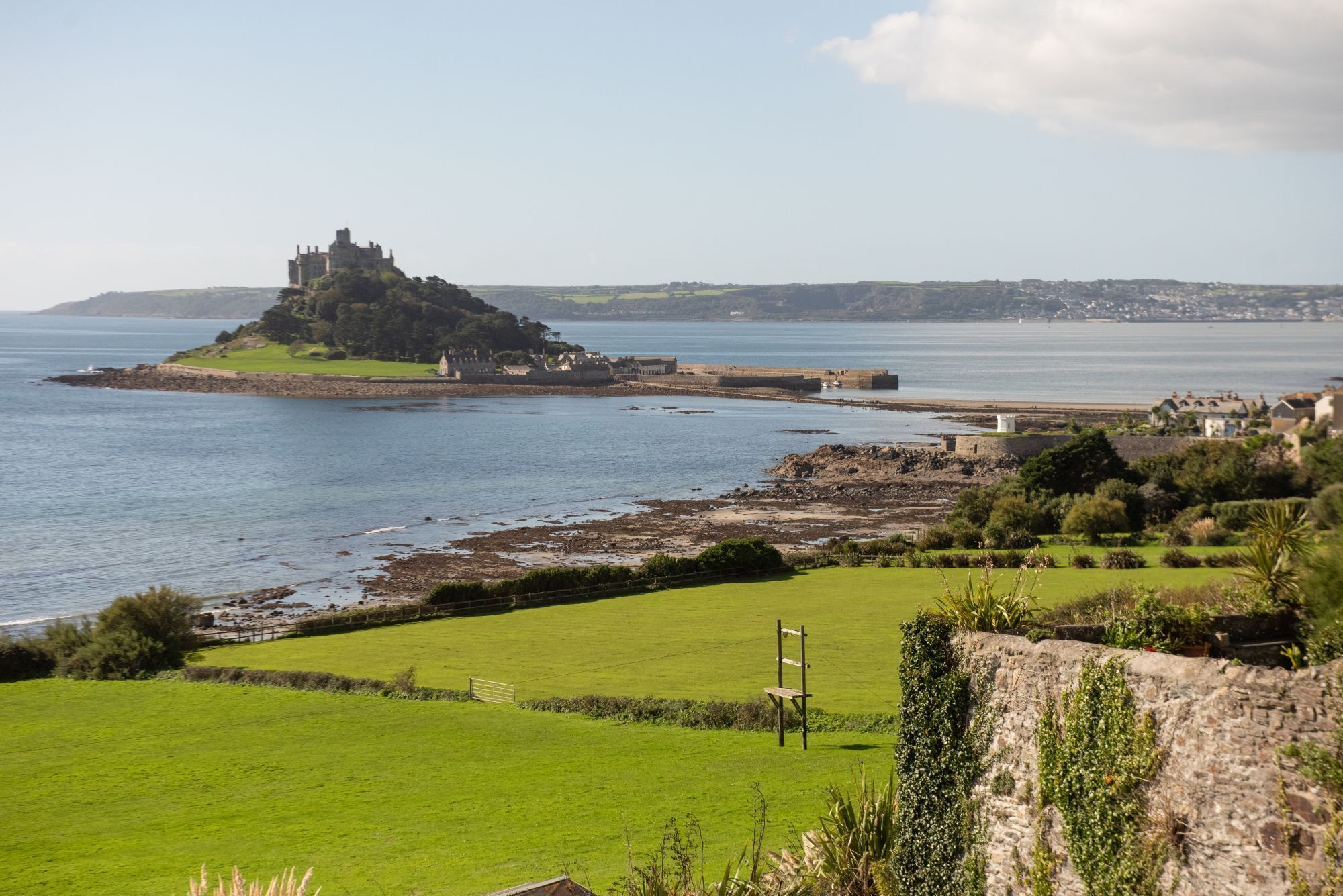 A coastal landscape with a historic castle on a small island, a grassy field, and a stone wall.