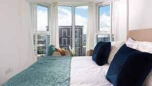 Ein Schlafzimmer mit großem Fenster, einem Bett mit blau-weißer Bettwäsche und Blick auf Gebäude.