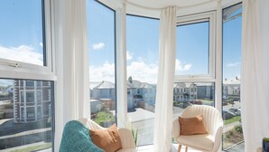 Ein Eckzimmer mit großen Fenstern, einem weißen Sofa, einem Sessel und Blick auf eine Wohngegend.