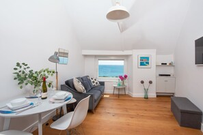 Ein modernes Wohnzimmer mit grauem Sofa, einem Esstisch mit Stühlen, einem kleinen Beistelltisch mit Lampe und Meerblick durch ein Fenster.