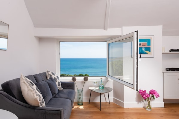 Ein modernes Wohnzimmer mit einem grauen Sofa, einem kleinen Tisch mit Getränken, einer Vase mit Blumen und einem großen Fenster mit Blick auf das Meer.