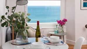 Una mesa de comedor dispuesta con platos y tazones blancos, y una botella de vino, con vista al mar a través de una ventana.