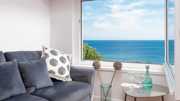 Un salón moderno con un sofá gris, un plaid blanco y gris con lunares, una mesita con dos vasos y una botella, y un ventanal grande con vistas al mar.