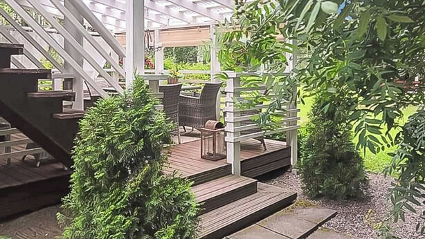 Ein überdachter Außenbereich mit Holzdeck, einer Pergola mit weißen Balken und einer Sitzgruppe mit Stühlen und Tisch.