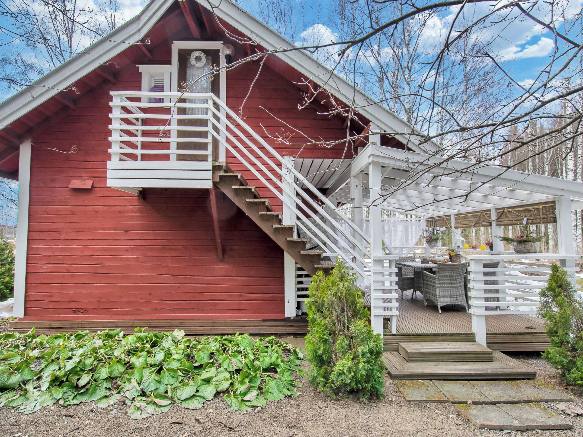 A red wooden cabin with a white staircase leading to a balcony, surrounded by greenery.