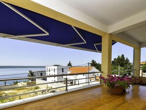 Un balcón con vistas al mar y edificios, una mesa de madera con una planta en maceta y un toldo azul.