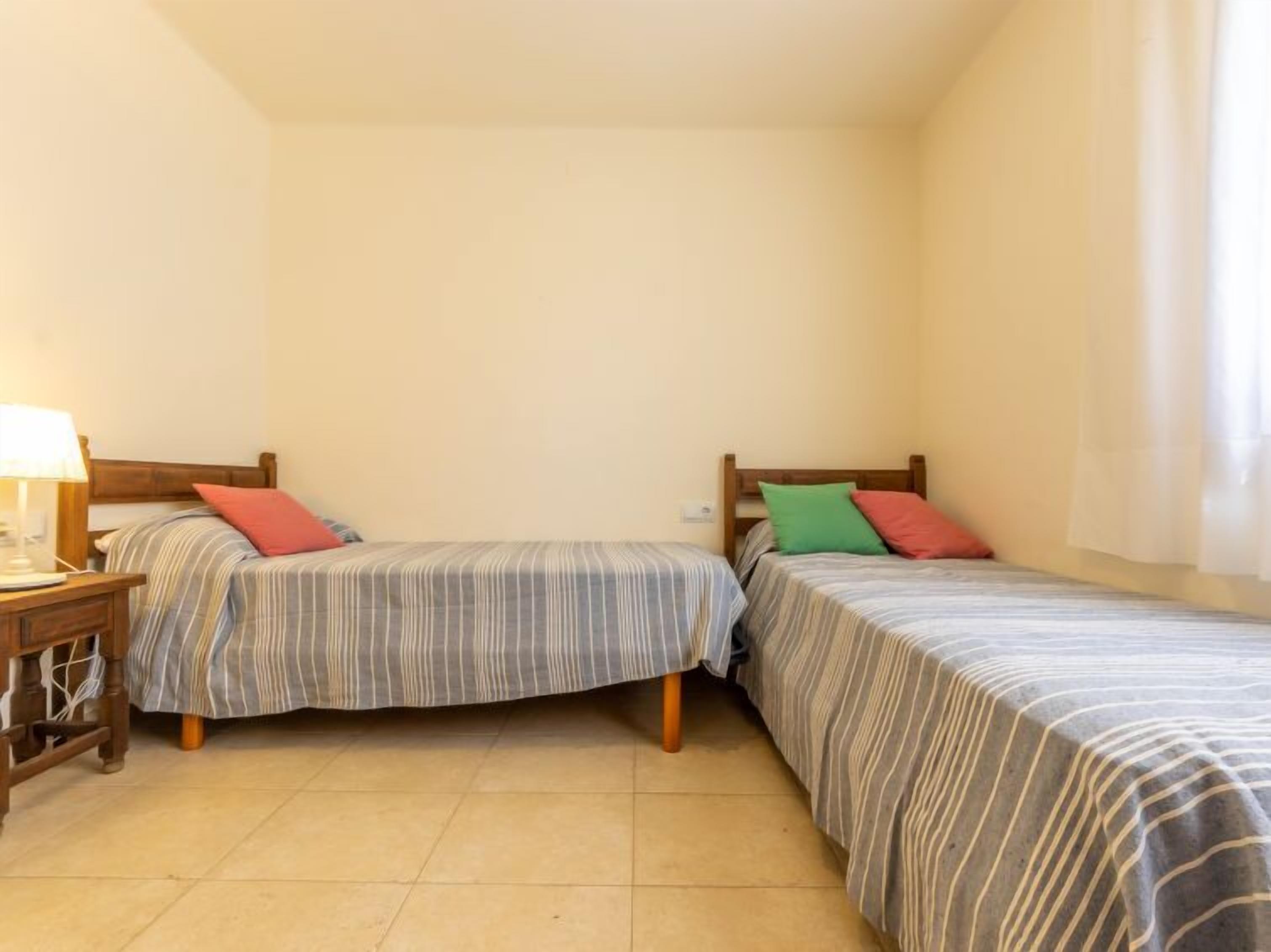 Une petite chambre propre, avec deux lits simples, une table de chevet avec une lampe et une fenêtre avec des rideaux.