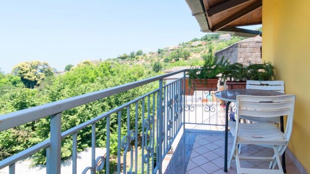Ein Balkon mit Tisch und Stühlen, der einen Blick auf eine Hügelkuppe mit Bäumen und Gebäuden freigibt.