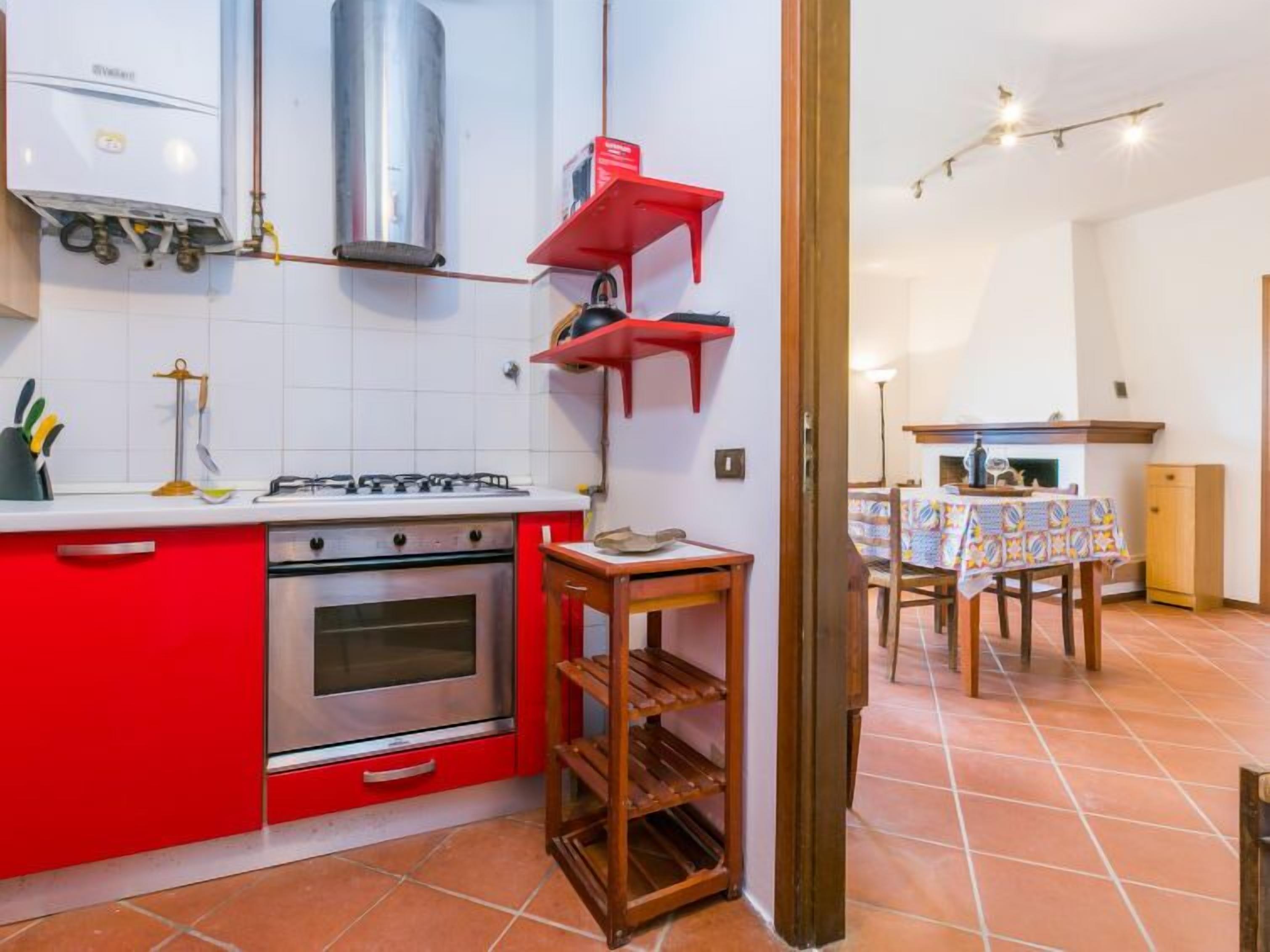 Een compacte keuken met een gasfornuis, oven en rode keukenkastjes. Er is een klein houten plankje en een betegelde spatwand.