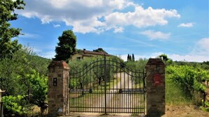 Una entrada portonada a una propiedad, con un portón de piedra, un camino que lleva a un edificio y un viñedo al fondo.