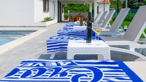 Un espace aménagé au bord de la piscine, avec des chaises longues et des parasols, une bouteille de vin et une table recouverte d’une nappe bleue.