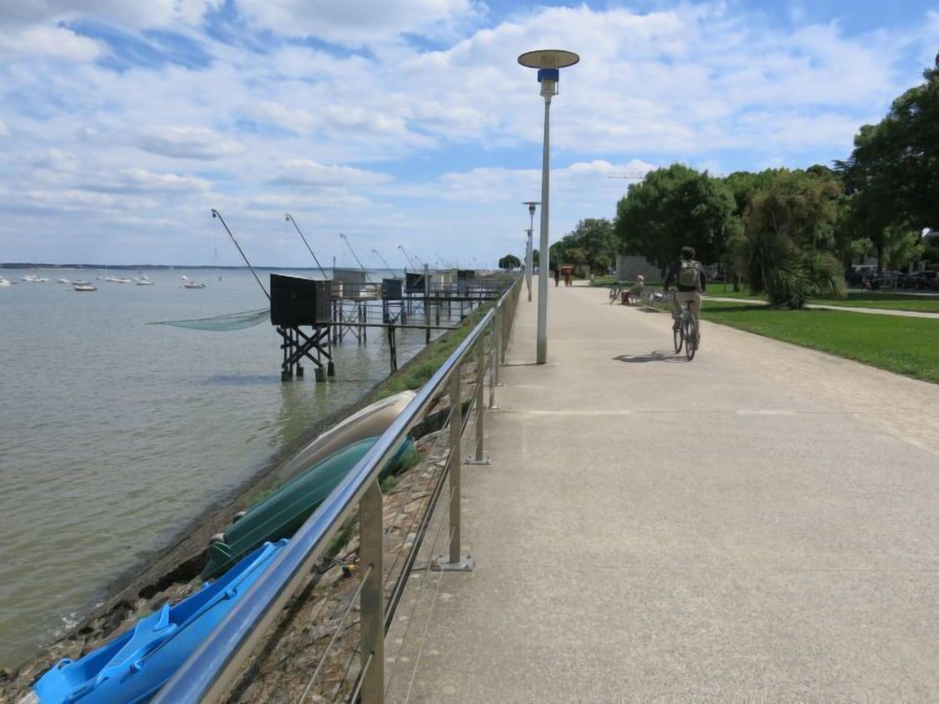 A waterfront walkway with a metal railing, a lamp post, and a cyclist.