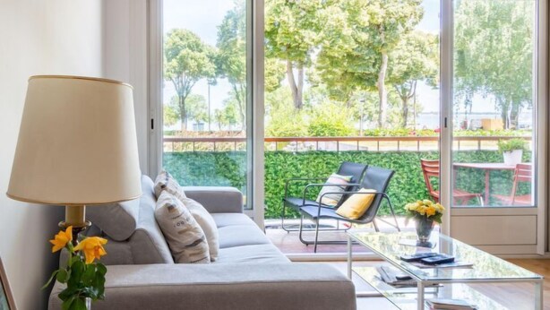 Una sala de estar moderna con un sofá, una mesa de centro de vidrio y una lámpara. Se ve un balcón con muebles de exterior y vegetación.