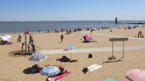 Una playa con gente tomando el sol y caminando, un cartel que dice "PLAGE DE VILLES-MARTIN", y un faro al fondo.