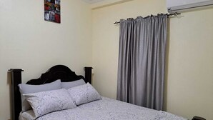 A single bed with a headboard, a bedside table, a grey curtain, and a wall-mounted air conditioning unit. - Cozy, comfortable, and beautiful apartment located in a safe gated community. (Georgetown)