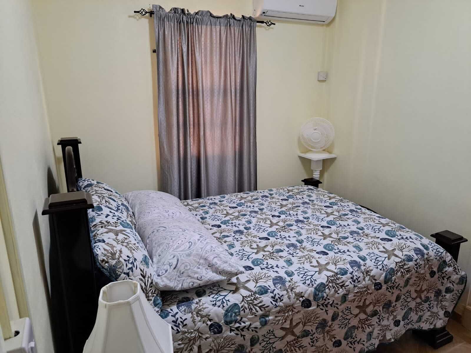 Een slaapkamer met een bed, nachtkastje, airconditioning aan de muur en een raam met gordijnen.