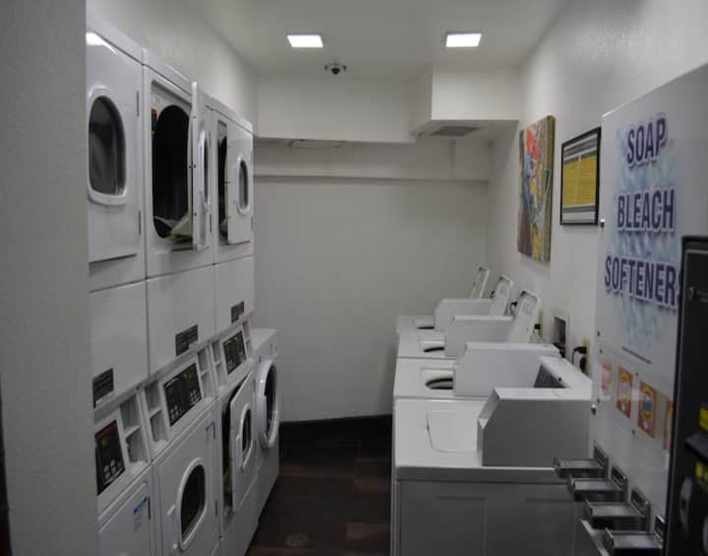Une buanderie équipée de plusieurs machines à laver et de plusieurs sèche-linge, d’un distributeur de savon et d’eau de Javel fixé au mur, et d’une caméra de surveillance installée au plafond.