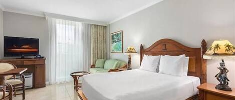 Habitación de hotel con una cama grande, un escritorio con televisor, una silla, una mesita pequeña y una mesita de noche con lámpara.