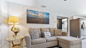 Uma sala de estar moderna com sofá em formato de L, uma mesa lateral de vidro com abajur e um quadro na parede retratando uma cena de praia.