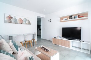 A modern living room with a flat-screen TV, a dining area with chairs, a coffee table, and a bookshelf with books.