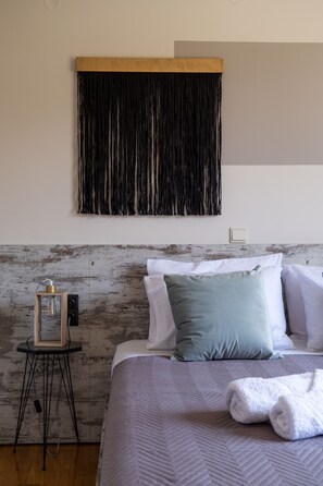 Un lit bien fait, avec une couverture grise, une serviette blanche et un oreiller vert. On y voit une tête de lit en bois, une tapisserie noire accrochée au mur et une petite table sur laquelle se trouve une lampe en verre.