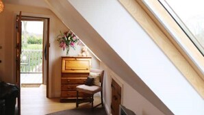 Ein Zimmer mit schrägem Dach, einem hölzernen Schminktisch, einem Stuhl und einer Vase mit Blumen.