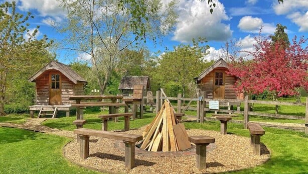 Ein rustikaler Campingplatz mit Holzhütten, Picknicktischen und einer zentralen Feuerstelle, umgeben von Kies.