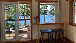 Una habitación con vistas a un lago y árboles, una mesa de madera con dos taburetes y una puerta corrediza de vidrio que da a una terraza exterior con muebles de mimbre.